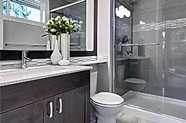 Bright new bathroom interior with glass walk in shower with grey tile surround, brown vanity cabinet topped with white counter and paired with mosaic tile backsplash.