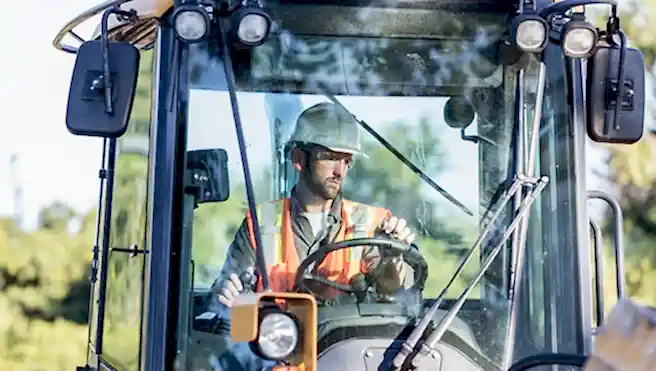 A mid adult man in his 30s working at a construction site, operating a backhoe. He is wearing a hardhat, safety vest and protective eyewear.